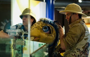 Two people in explorer hats handling a large, blue-eyed dinosaur puppet.