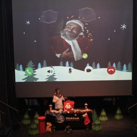 A member of the learning team standing next to a table of Christmas items. She is in front of a large screen with Santa on it.
