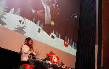 A member of the learning team wearing an elf hat, giving a presentation. Behind is a Christmas scene on the screen.