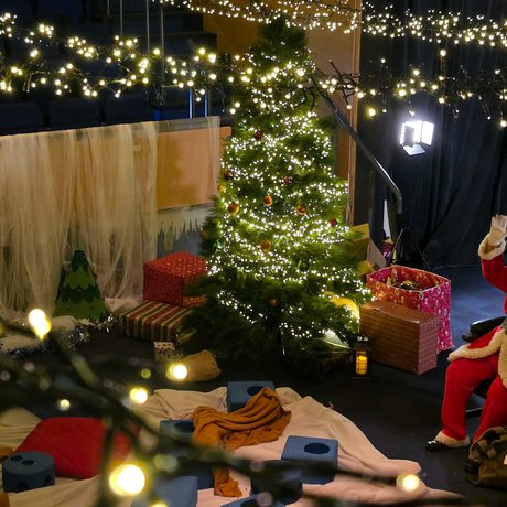 Santa Claus waving next to a decorated Christmas tree surrounded by gifts and festive lights.