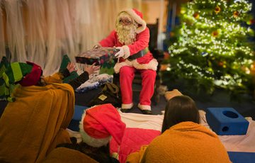 Santa Claus hands a gift to an excited person near a decorated Christmas tree.