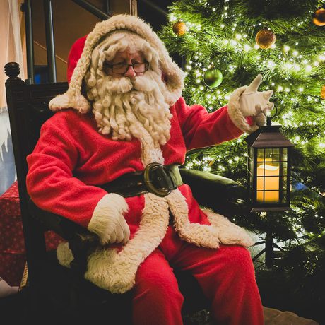 Santa Claus in classic red suit holding lantern, sitting beside a decorated, glowing Christmas tree.