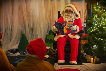 Santa Claus reads a Christmas story to children beside a decorated tree with lights.