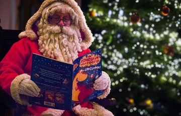 Santa Claus reads a colourful children's book by a decorated, glowing Christmas tree.