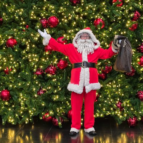Santa standing in between two large nut cracker decorations and in front of a large christmas tree decorated with red baubles and lights.