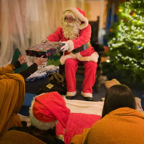 Santa Claus hands a gift to an excited person near a decorated Christmas tree.