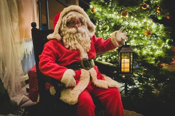 Santa Claus in classic red suit holding lantern, sitting beside a decorated, glowing Christmas tree.