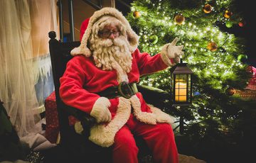 Santa Claus in classic red suit holding lantern, sitting beside a decorated, glowing Christmas tree.