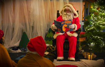 Santa Claus reads a Christmas story to children beside a decorated tree with lights.