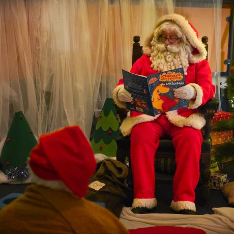 Santa Claus reads a Christmas story to children beside a decorated tree with lights.