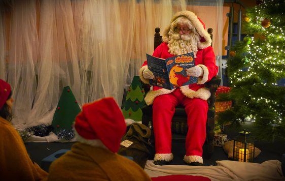 Santa Claus reads a Christmas story to children beside a decorated tree with lights.