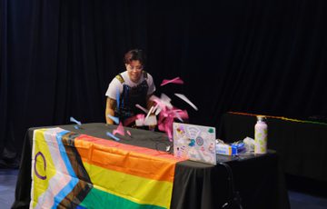 Person joyfully performing an experiments with pink papers flying off above a rainbow pride flag-covered table.