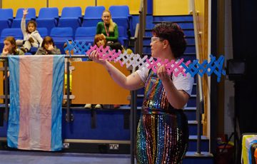 Person in rainbow outfit performs with colorful prop while audience watches, trans pride flag displayed.