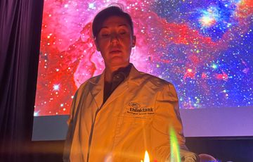 A staff member in a lab coat doing a science experiment with bright colour flames on a table in front of them.