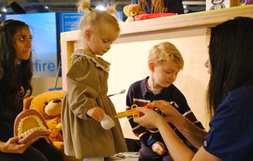 A toddler and child play with toy doctors and dentist equipment guided by adults.