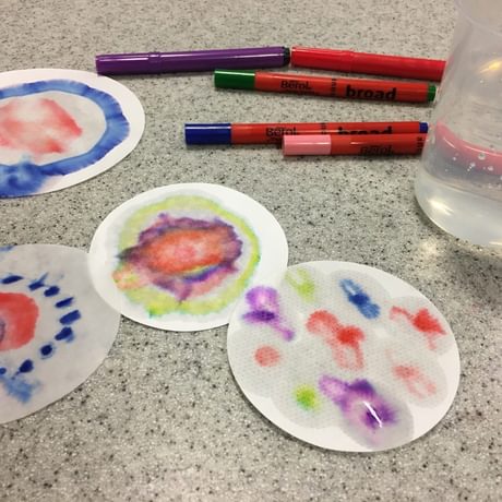 Objects on a table inducing: Filter paper with coloured patterns on it, pens and a glass filled with liquid.