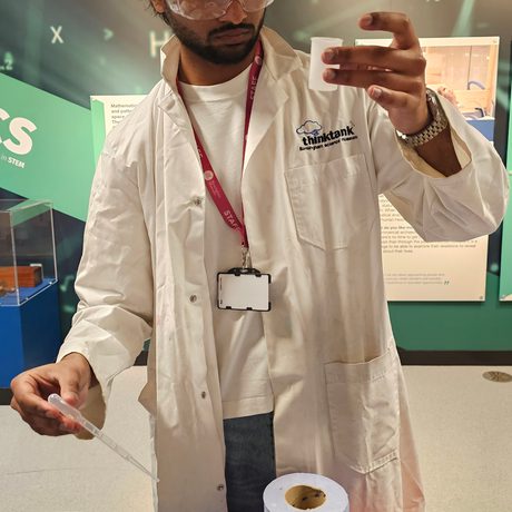 A staff member wearing googles and lab coat holding a canister and other equipment on a table in front.