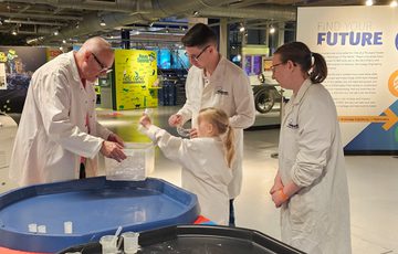A group of three adults and a child wearing lab coats getting a canister out of a plastic box.