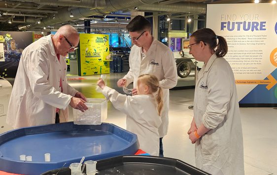 A group of three adults and a child wearing lab coats getting a canister out of a plastic box.