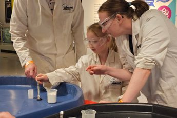 And adult and a child wearing lab coats putting liquid into a canister.