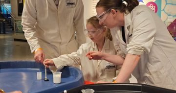 And adult and a child wearing lab coats putting liquid into a canister.