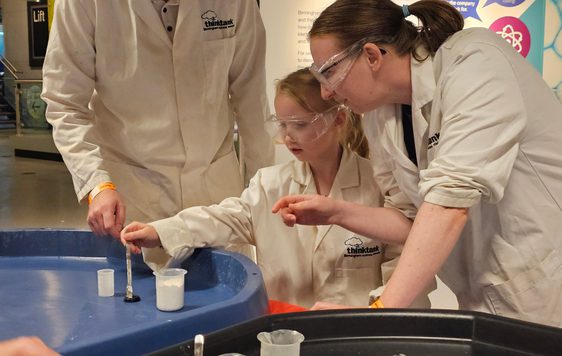 And adult and a child wearing lab coats putting liquid into a canister.