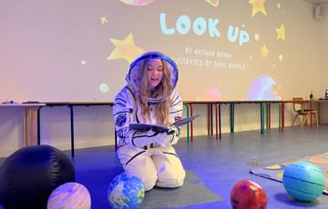 A person dressed as an astronaut sitting on the floor reading a book with model planets around them.