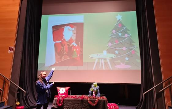 A member of the learning team standing next to a table of Christmas items. She is in front of a large screen with Santa on it.