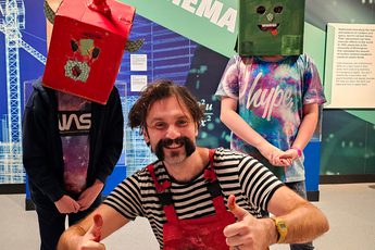 A person crouching holding their thumbs up next to two children wearing cardboard box head sculptures.