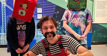 A person crouching holding their thumbs up next to two children wearing cardboard box head sculptures.