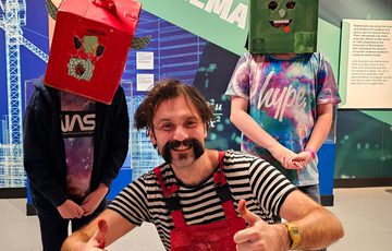 A person crouching holding their thumbs up next to two children wearing cardboard box head sculptures.
