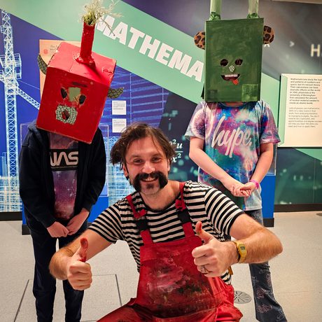 A person crouching holding their thumbs up next to two children wearing cardboard box head sculptures.