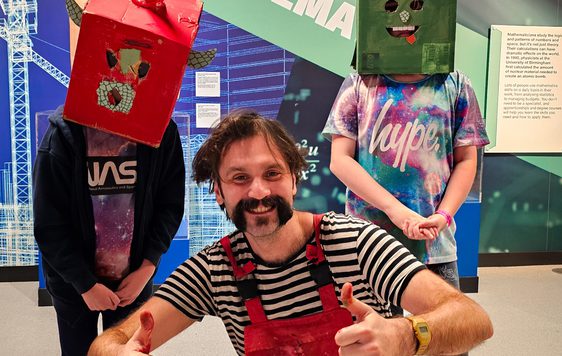 A person crouching holding their thumbs up next to two children wearing cardboard box head sculptures.