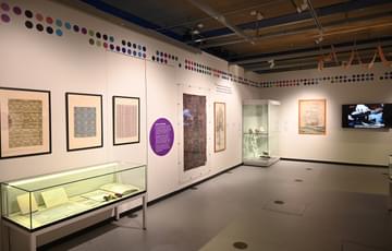 An exhibition wall featuring patterns and designs on the wall along with display cases of small paper objects.