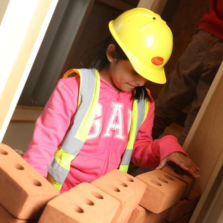 A child wearing a toy hard hat while building a toy foam brick wall.