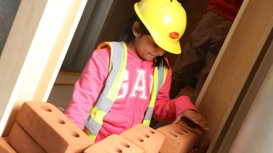 A child wearing a toy hard hat while building a toy foam brick wall.