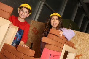 An adult and child smiling while wearing toy hard hats while building walls with foam bricks.