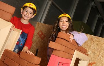 An adult and child smiling while wearing toy hard hats while building walls with foam bricks.