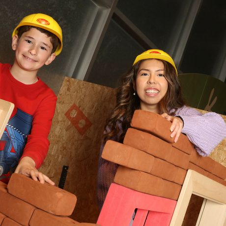 An adult and child smiling while wearing toy hard hats while building walls with foam bricks.