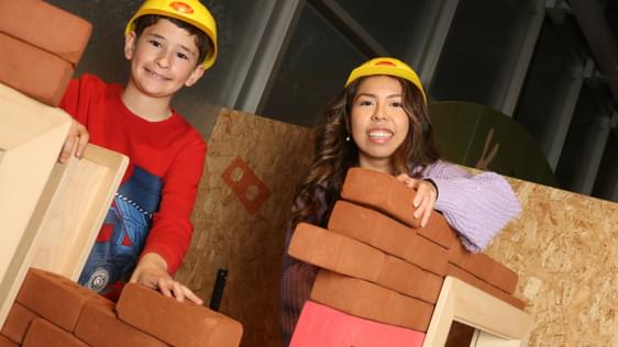 An adult and child smiling while wearing toy hard hats while building walls with foam bricks.