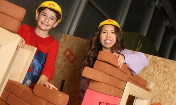 An adult and child smiling while wearing toy hard hats while building walls with foam bricks.