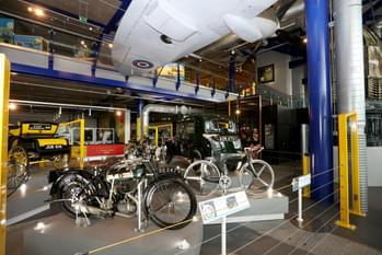 Exhibition hall with vintage motorcyles, cars and other bikes. A Spitfire plane is hanging above.