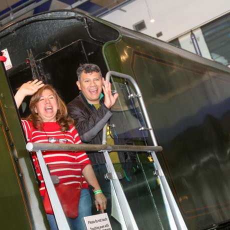 Two adults waving from a steam train that is on display.