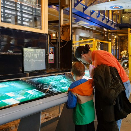 A woman and child explore an interactive exhibit inside a museum with vintage vehicles.