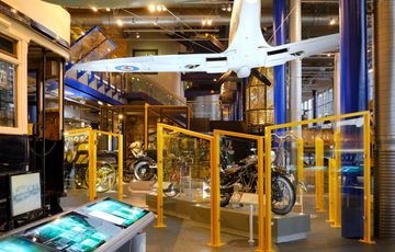 Historic motorcycles, vintage vehicles, and a suspended Spitfire airplane displayed in a museum setting.