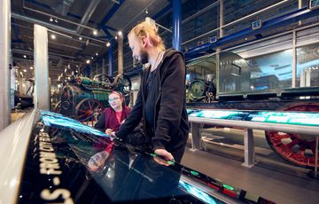 Two people interact with large displays inside a museum with vintage machinery.