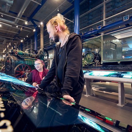 Two people interact with large displays inside a museum with vintage machinery.