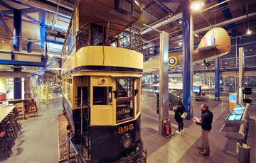Historic yellow and black tram displayed indoors with visitors observing exhibits nearby.