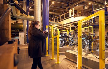 A woman photographs vintage bicycles displayed behind yellow barriers in an industrial museum gallery.