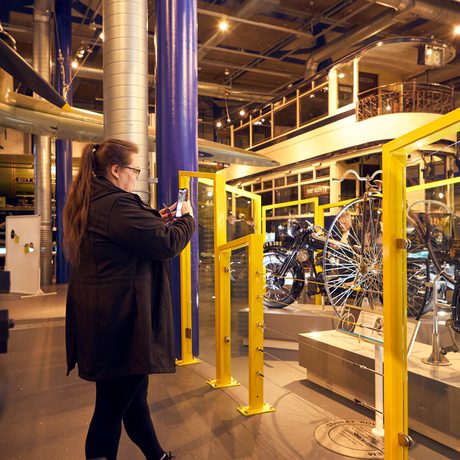 A woman photographs vintage bicycles displayed behind yellow barriers in an industrial museum gallery.